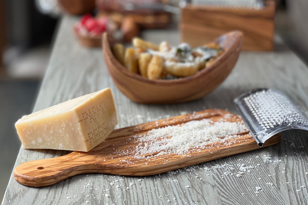 Flat wooden cheese grater with handle holding grated Parmesan cheese, displayed with wedge of cheese.