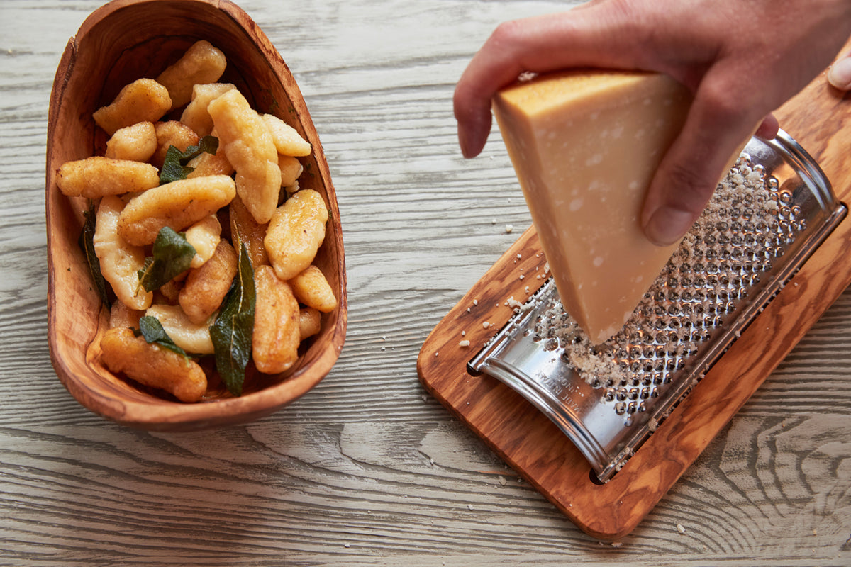 Flat olivewood grater with stainless steel grating surface, held over block of hard cheese, with fried snacks in wooden bowl.