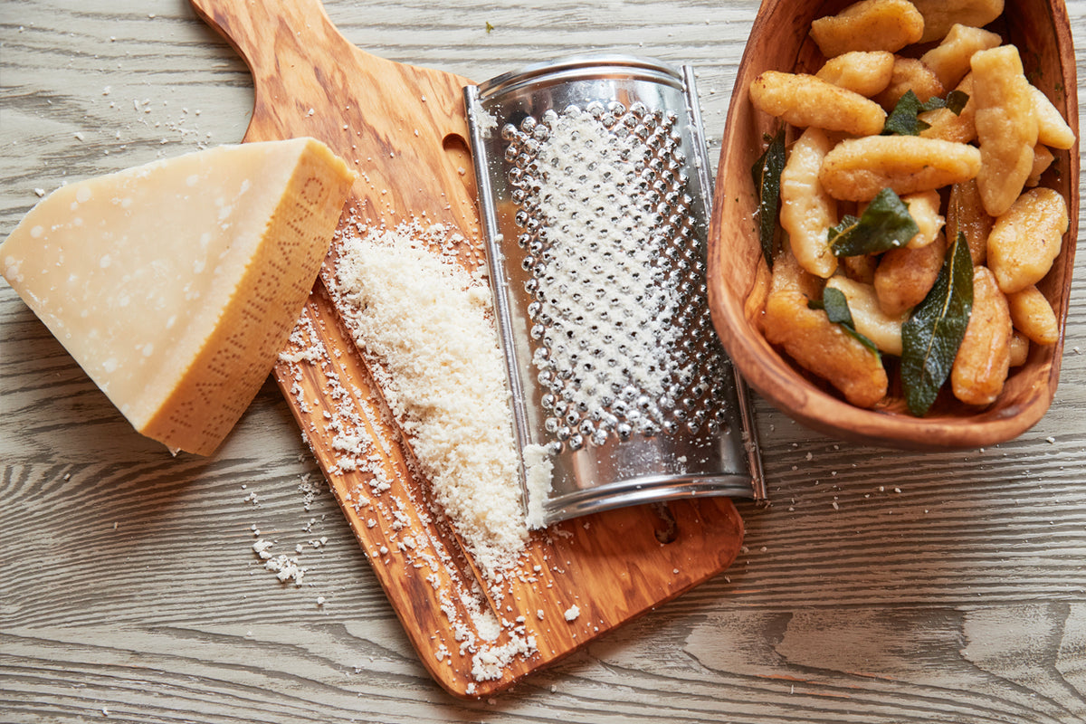 Stainless steel flat grater with olivewood handle on wooden surface, surrounded by Parmesan cheese and fried potatoes.