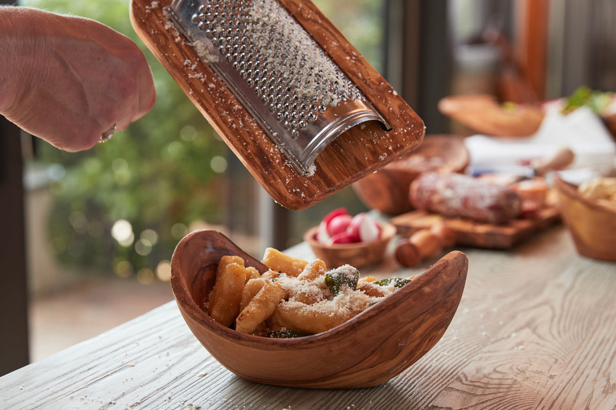 Wooden cheese grater with metal grating surface held above matching wooden bowl containing pasta with grated cheese.