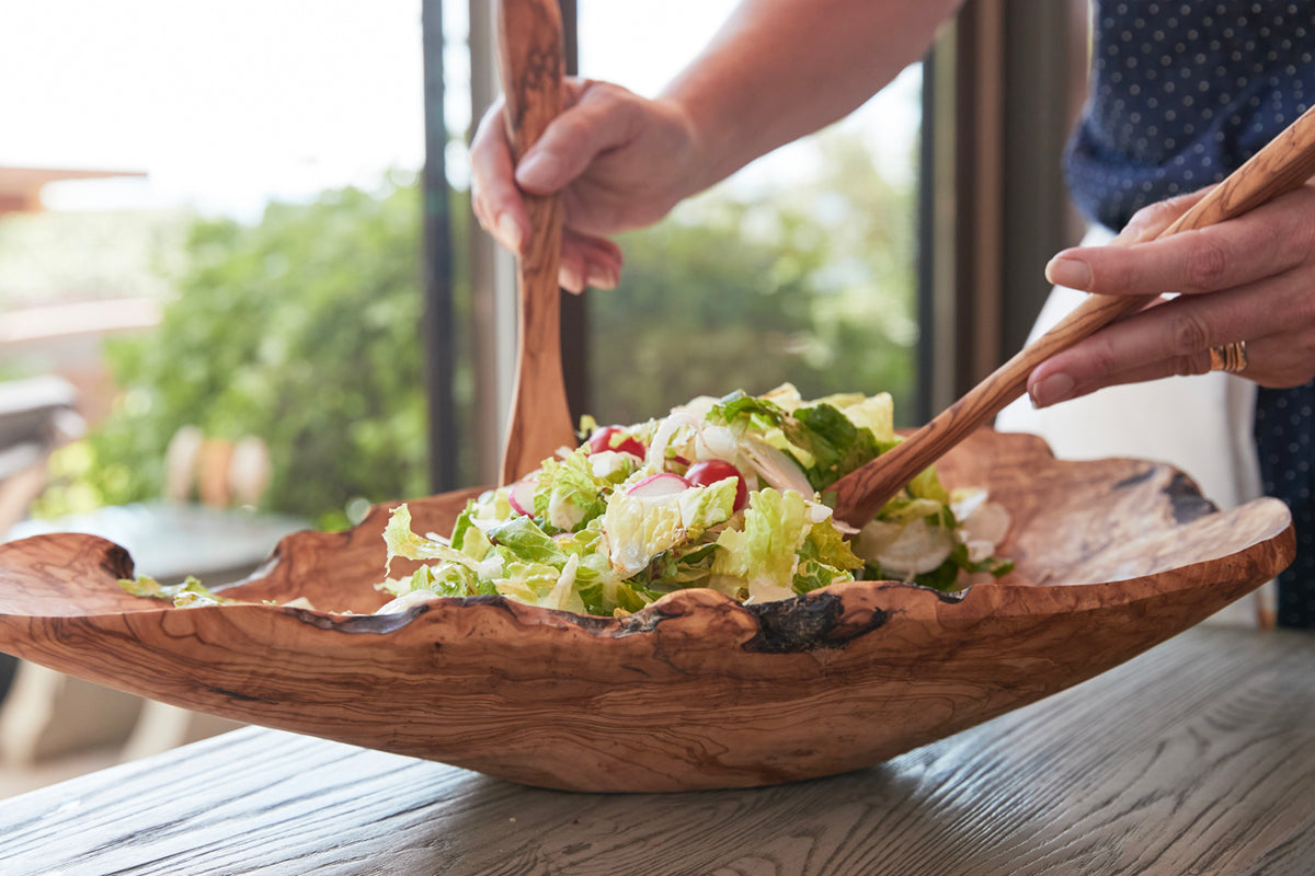 Hands tossing salad with wooden utensils in a carved olivewood bowl with natural root edges.