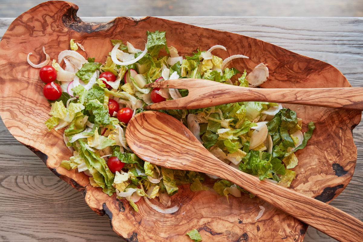 Rustic wooden salad bowl with organic edges containing mixed greens, red tomatoes, and onions with two wooden serving utensil