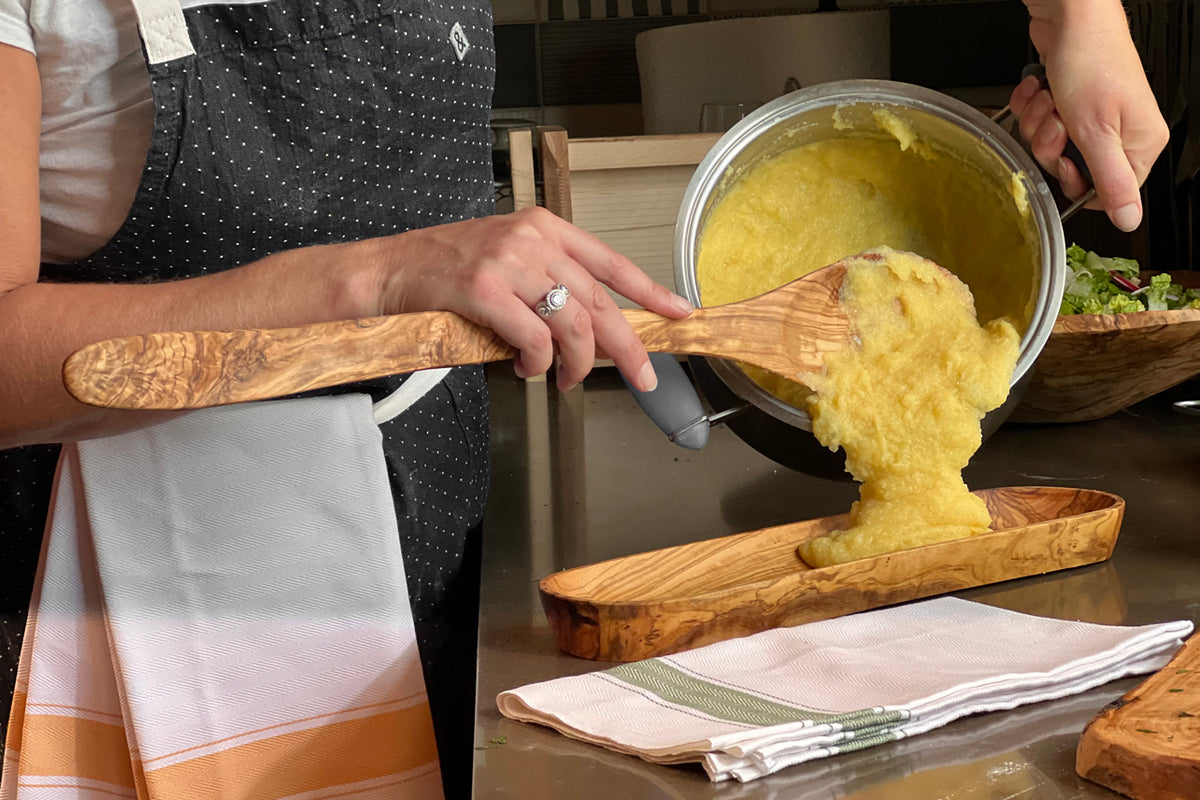 Wooden spoon with wide, flat paddle stirring yellow polenta in metal pot over wooden serving board.
