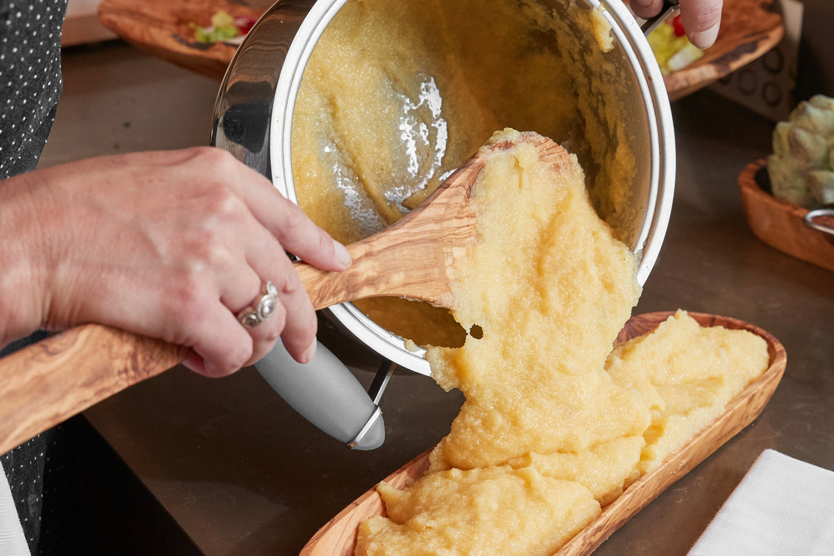 Wooden spoon with flat blade stirring creamy polenta in white pot over wooden serving board.