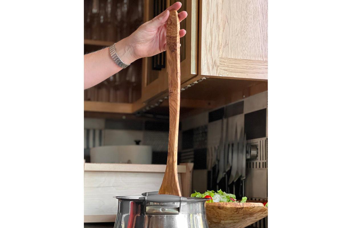 Hand holding long wooden spoon with rounded bowl over pot in kitchen setting.
