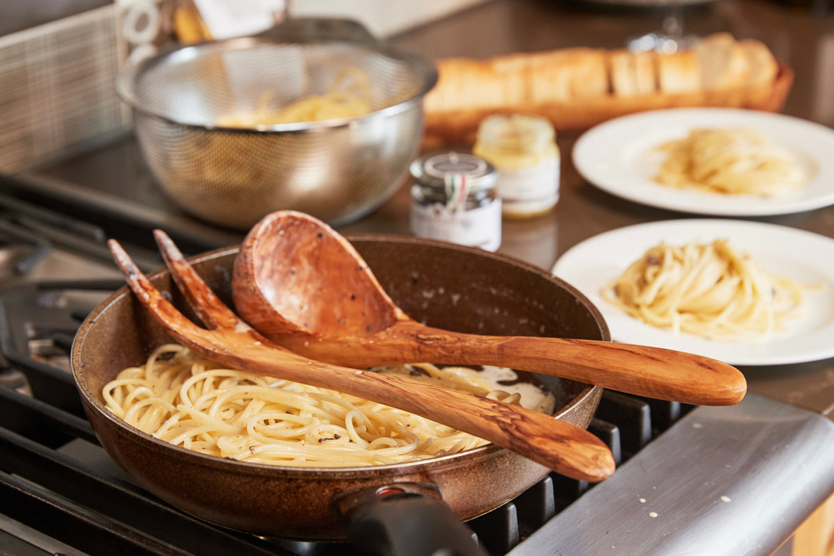 Wooden ladle with long handle resting in pasta pan on stovetop with cooked noodles.
