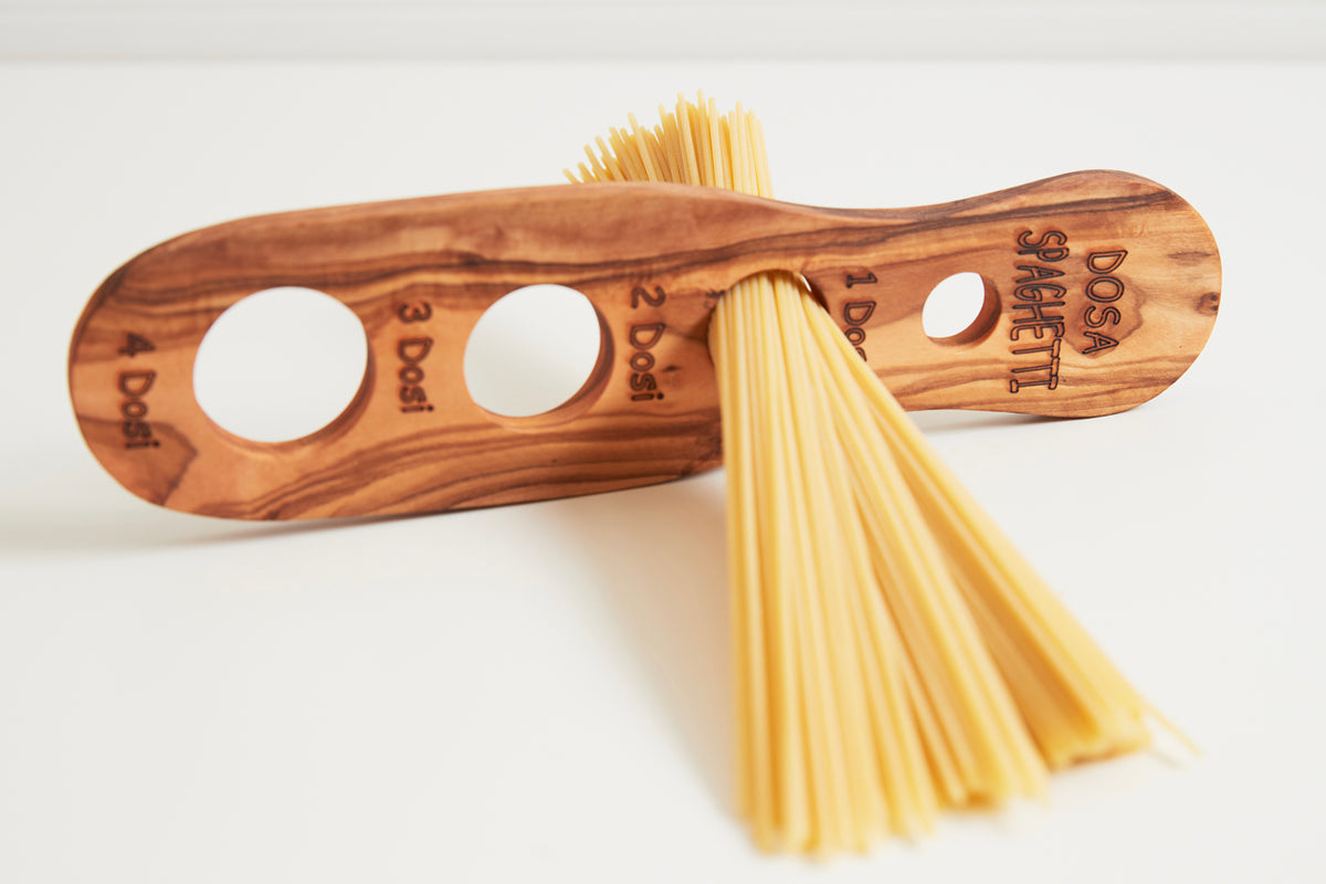 Wooden spaghetti measurer with four circular holes and engraved portion markings, holding dried spaghetti noodles.