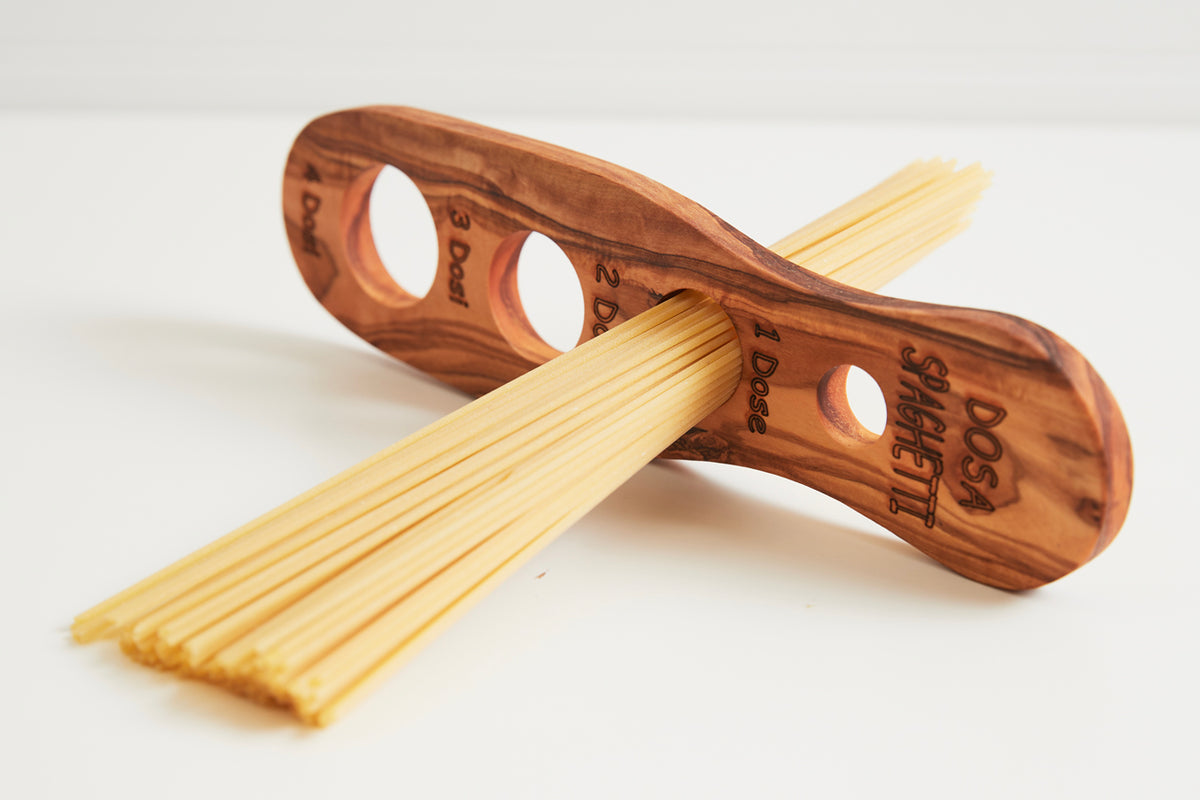 Wooden spaghetti measurer with multiple circular holes and engraved portion markings, holding dry pasta strands.