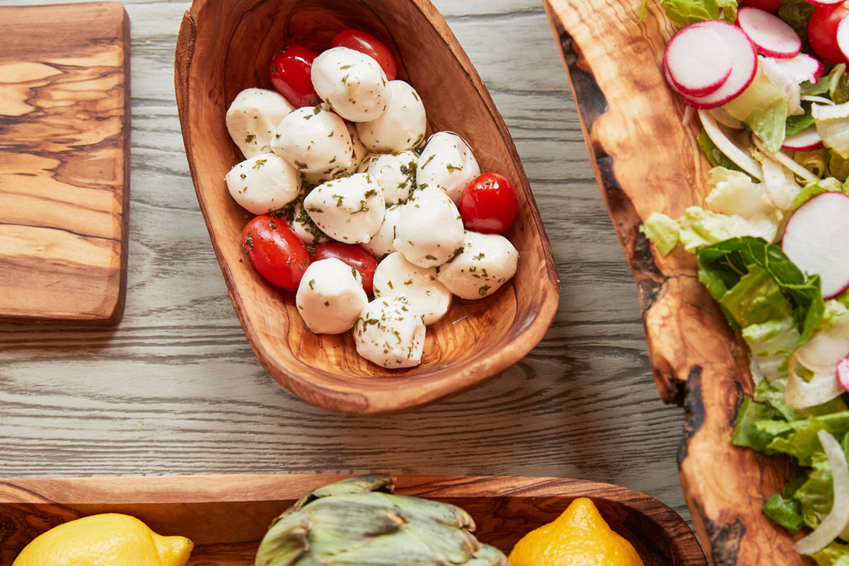 Oval olivewood serving bowl filled with mozzarella balls, cherry tomatoes, and herbs on gray wood surface.