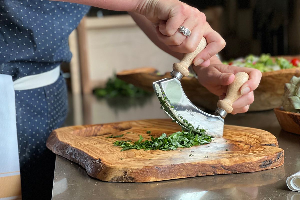 Hand using curved knife with wooden handle to chop herbs on large olivewood cutting board.