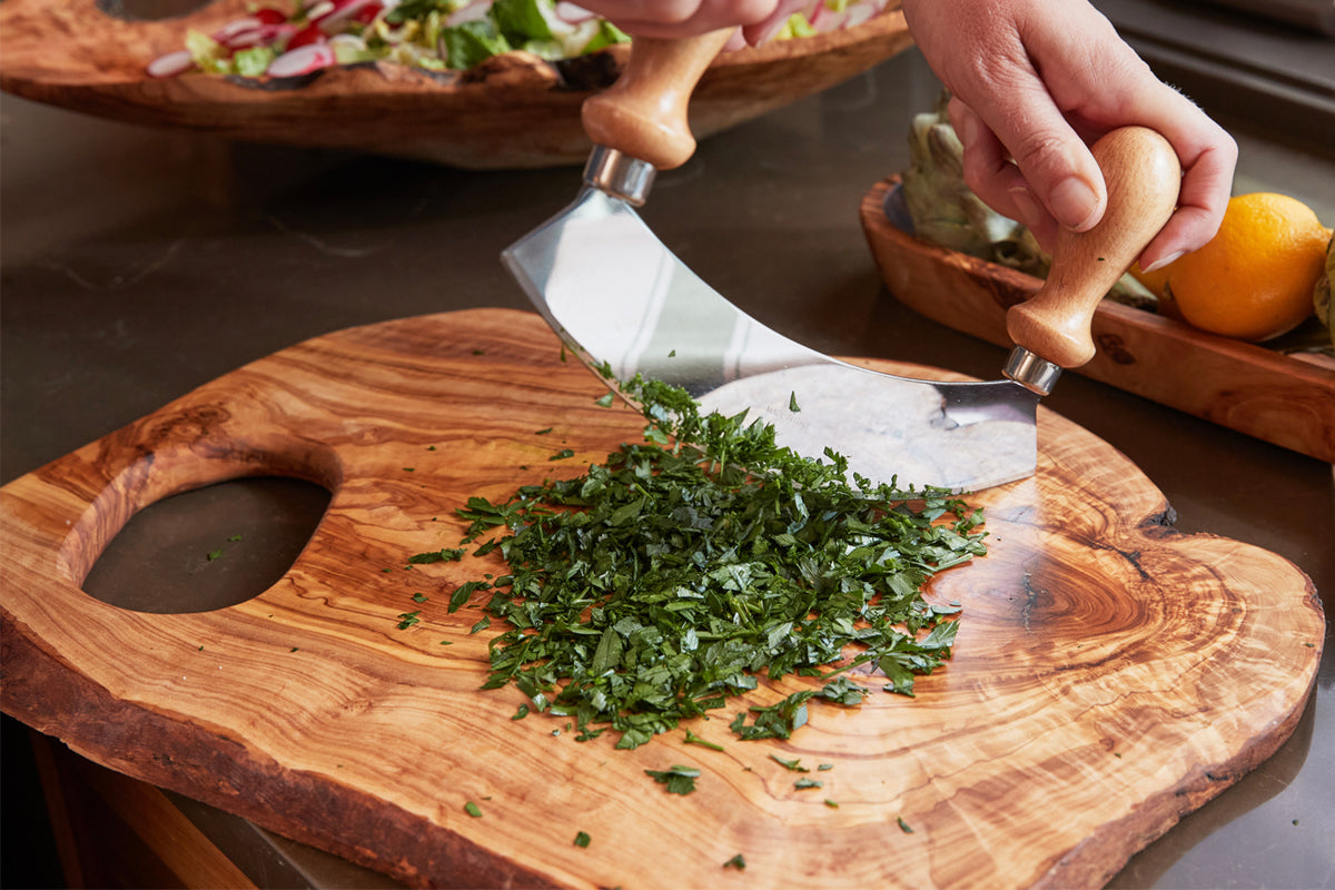 Large olivewood cutting board with handle featuring chopped herbs and knife, surrounded by additional boards and fresh ingred