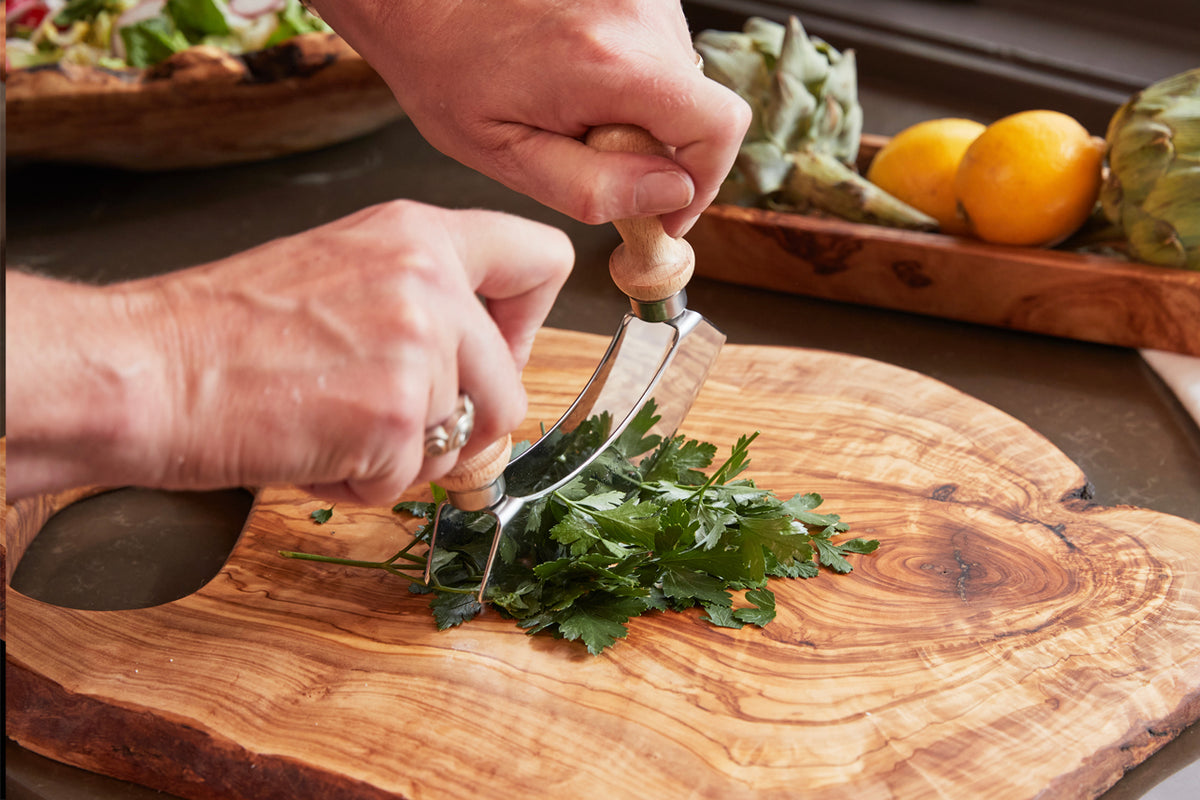 Large olive wood cutting board with natural grain patterns, being used to chop fresh herbs with a curved mezzaluna knife.