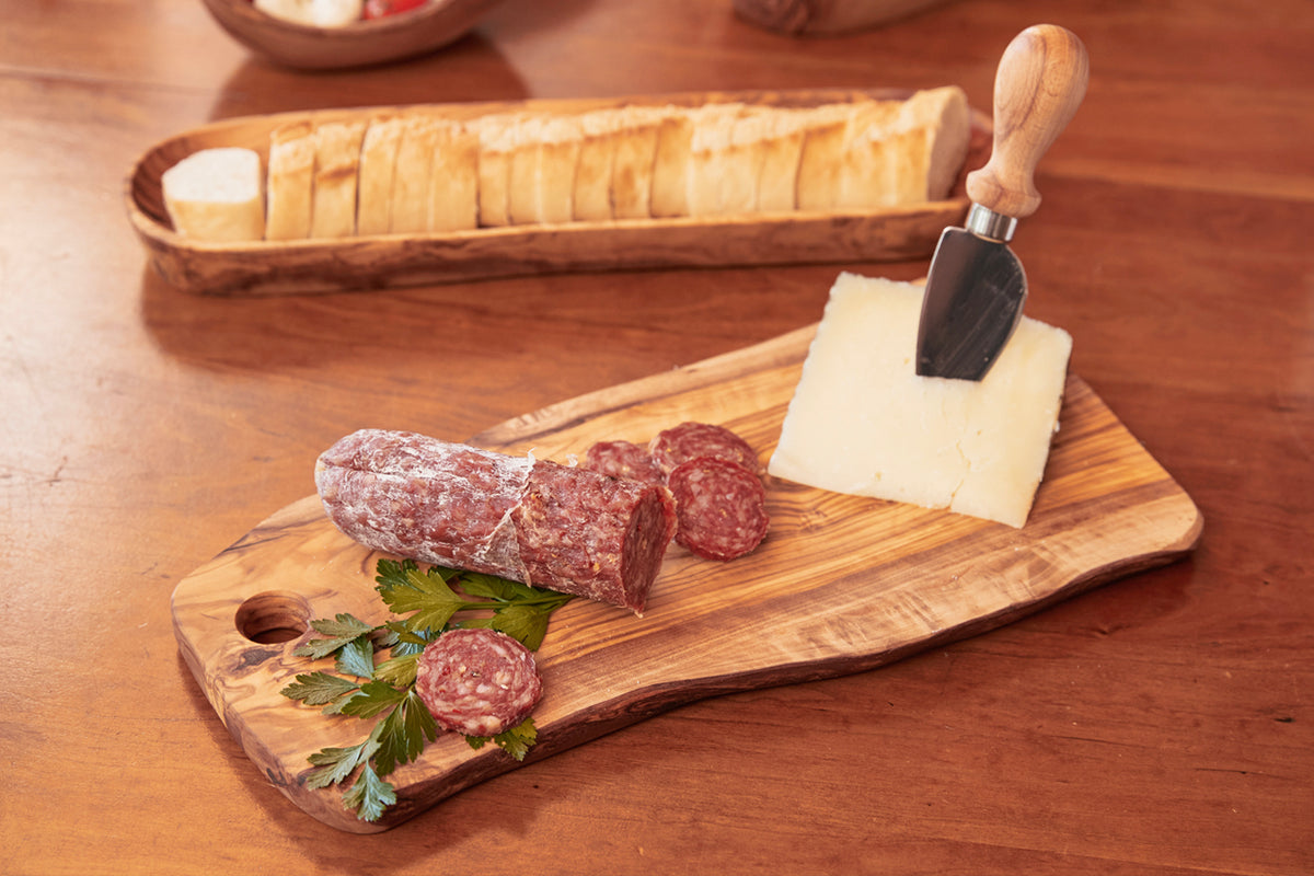 Olivewood charcuterie board with hole, sliced salami, cheese, knife, and bread basket.