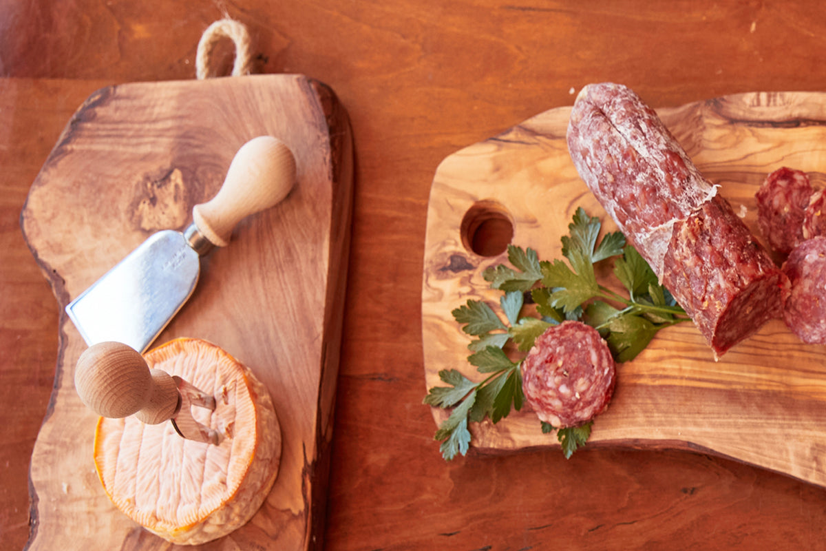 Olivewood charcuterie board with rope handle, cheese knife, sliced salami, and orange garnish.