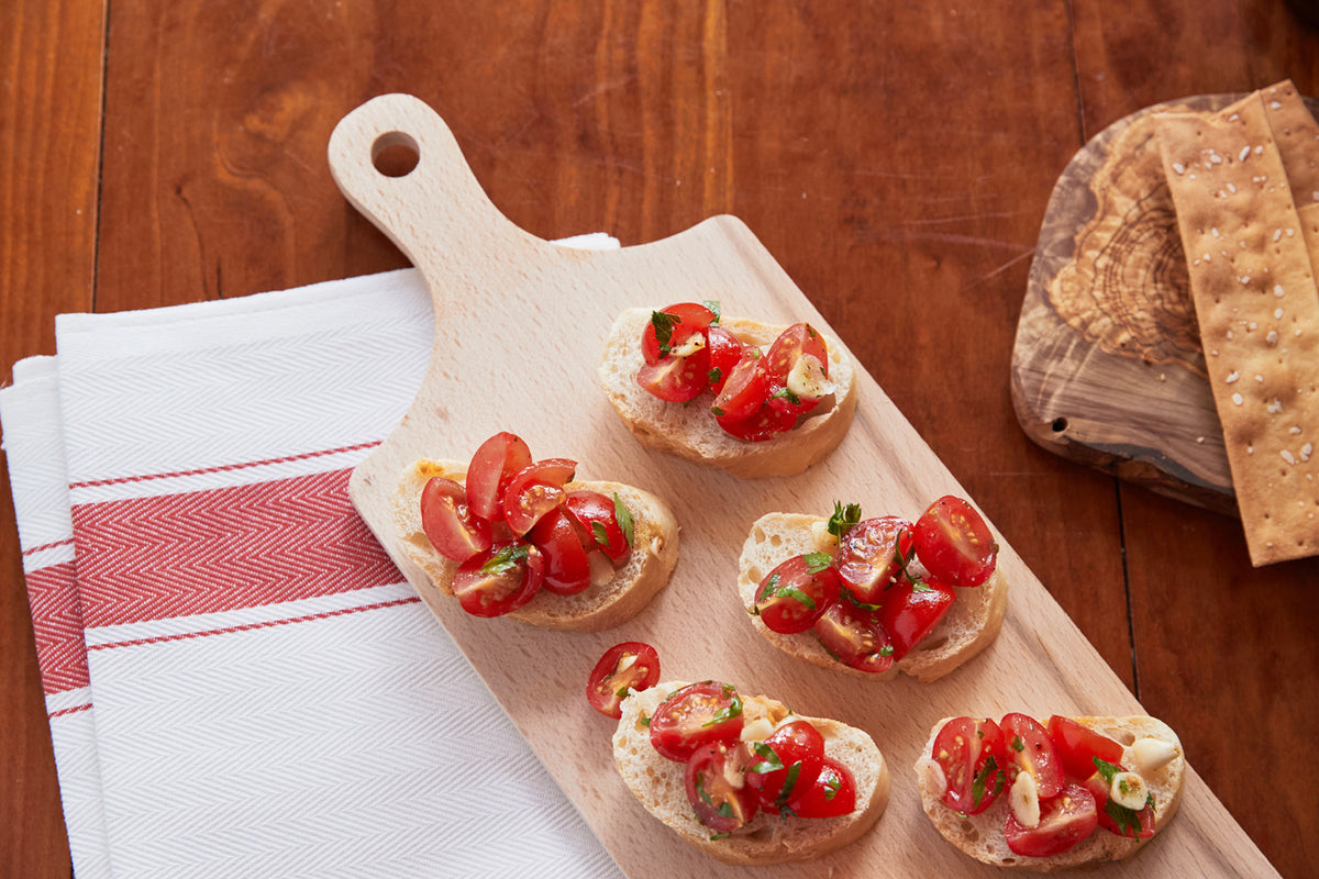 Light wood paddle board with handle holding five bruschetta pieces topped with tomatoes and herbs on wooden surface.