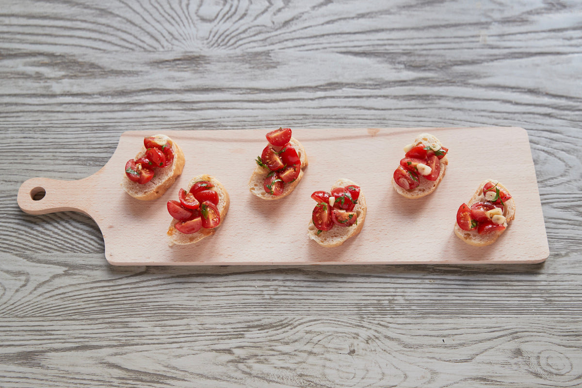 Light wooden paddle board with handle displaying six pieces of bruschetta topped with tomatoes and herbs.