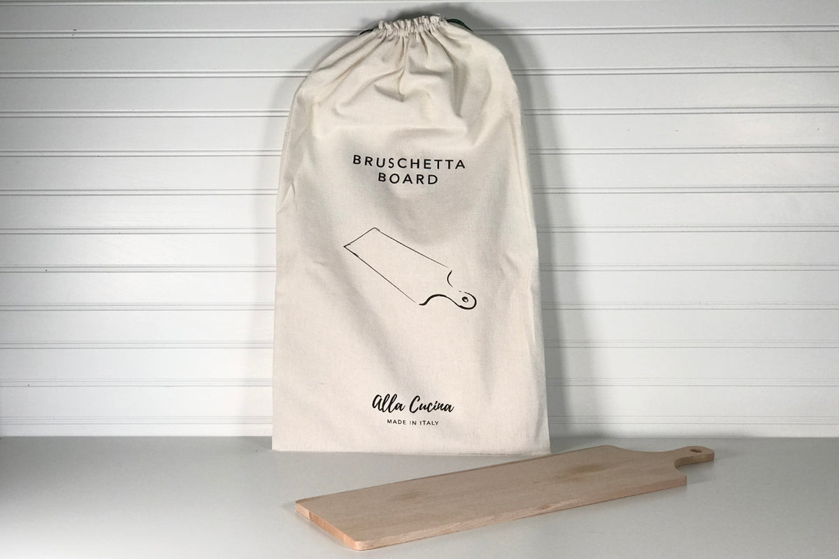 Light wood bruschetta board with handle next to cream cotton drawstring storage bag labeled "Bruschetta Board."