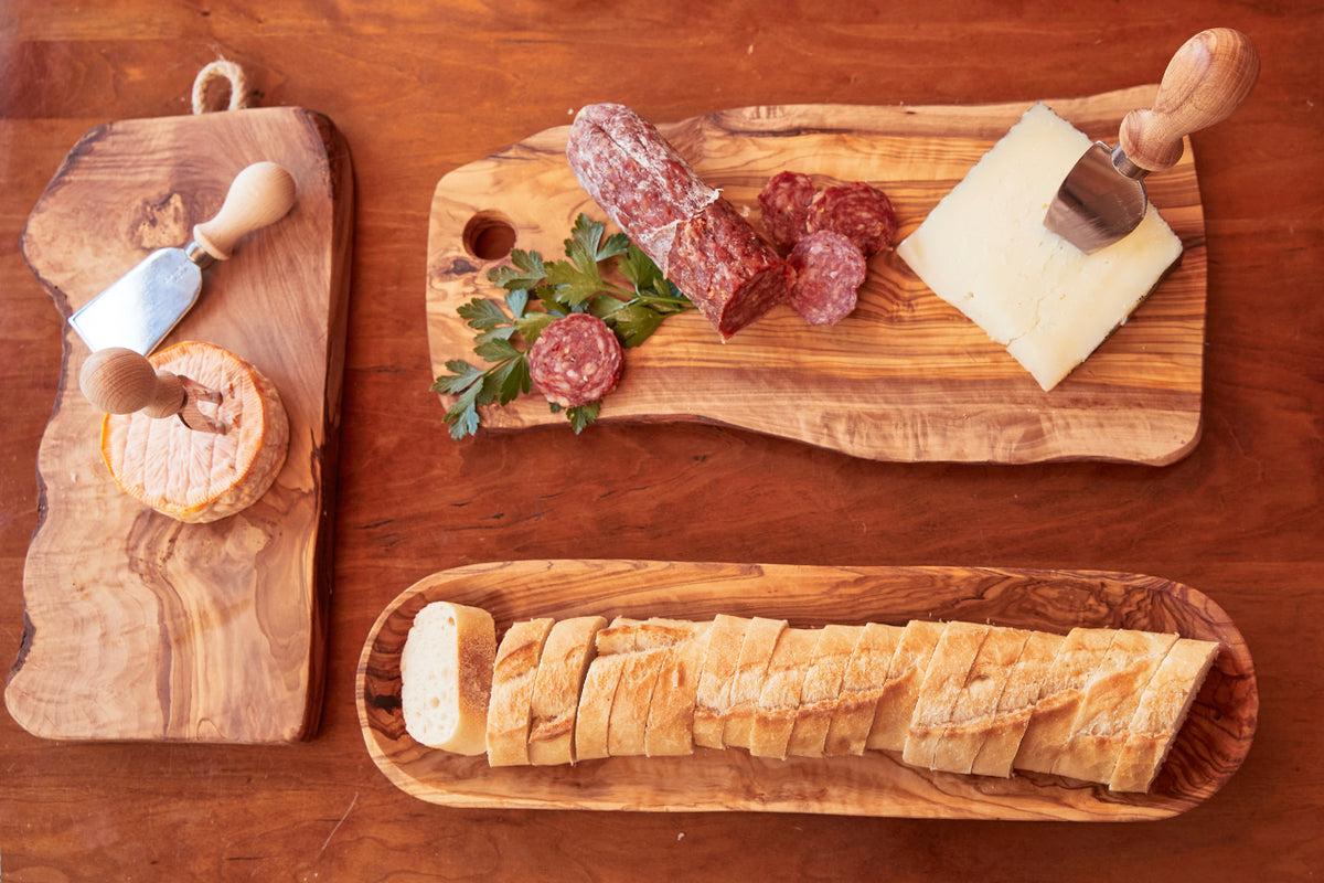 Three cheese knives with wooden handles displayed on wooden boards with sliced cheese and cured meat.
