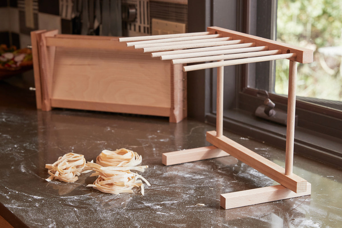 Wooden pasta drying rack with multiple horizontal rods and a collapsible fabric bin, displayed with fresh pasta nests on a co