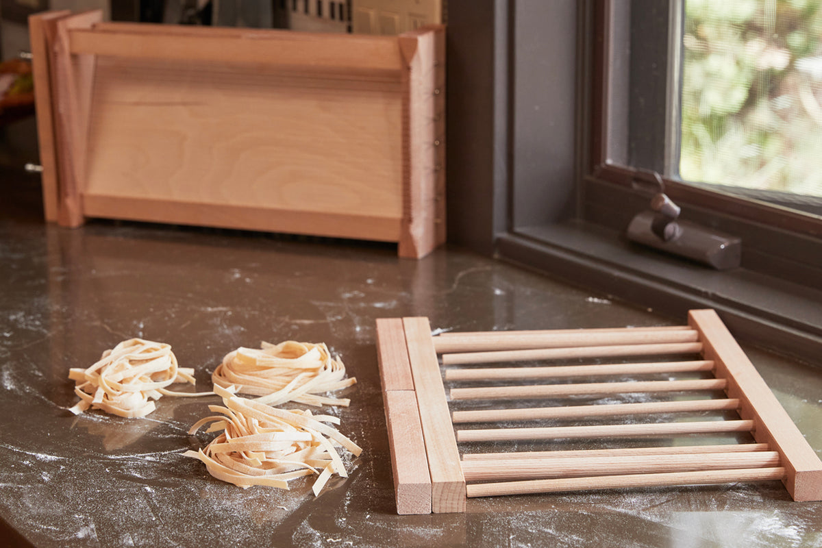 Wooden pasta drying rack with parallel rods and fresh pasta nests on a countertop, with a collapsed rack in the background.