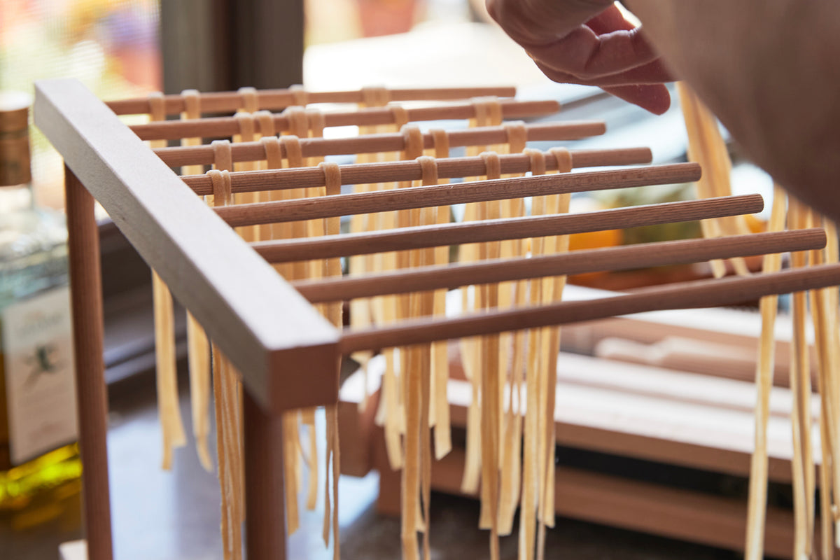 Wooden pasta drying rack with multiple horizontal rods holding fresh pasta strands, collapsible frame visible.