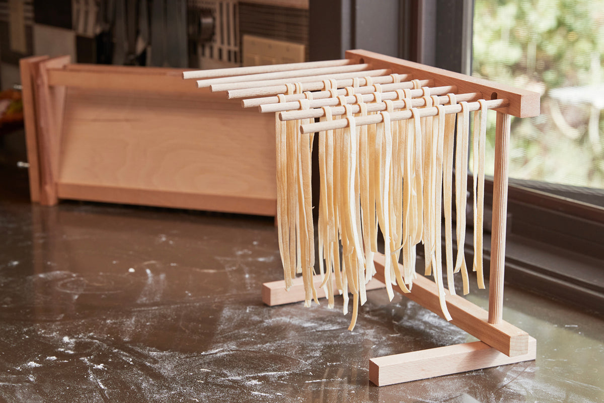 Wooden pasta drying rack with fresh pasta strands draped over multiple horizontal rods, alongside a folded collapsible tray.