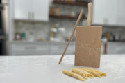Wooden pasta board with vertical ridges, two wooden rolling pins, and fresh garganelli pasta on marble countertop.
