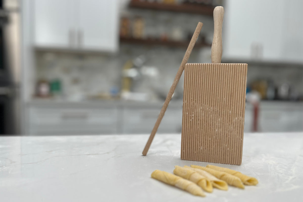 Wooden pasta board with vertical ridges, two wooden rolling pins, and fresh garganelli pasta on marble countertop.