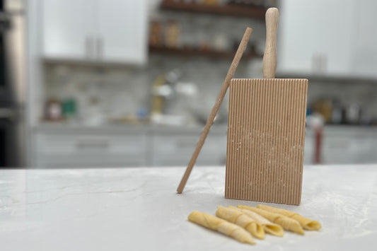 Wooden pasta board with vertical ridges, two wooden rolling pins, and fresh garganelli pasta on marble countertop.