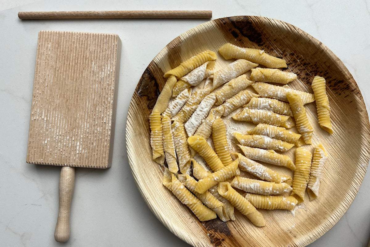 Wooden pasta board with ridged surface, rolling pin, and woven basket bowl filled with fresh gnocchi and garganelli pasta.