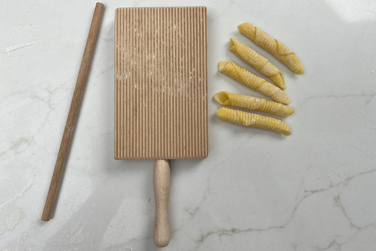 Wooden pasta board with ridged surface, rolling pin, and five yellow garganelli pasta shapes on marble background.