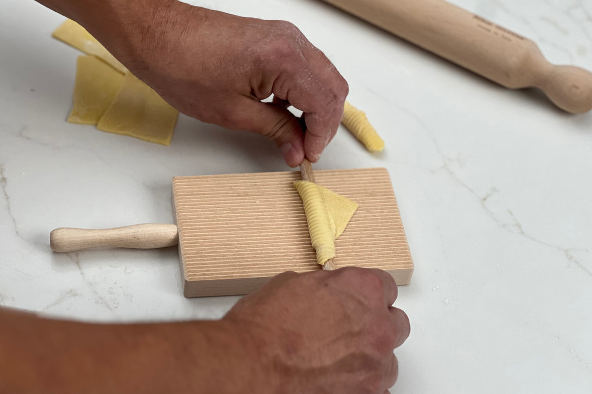 Wooden pasta board with ridged surface and handle being used to shape fresh yellow pasta with a rolling pin nearby.