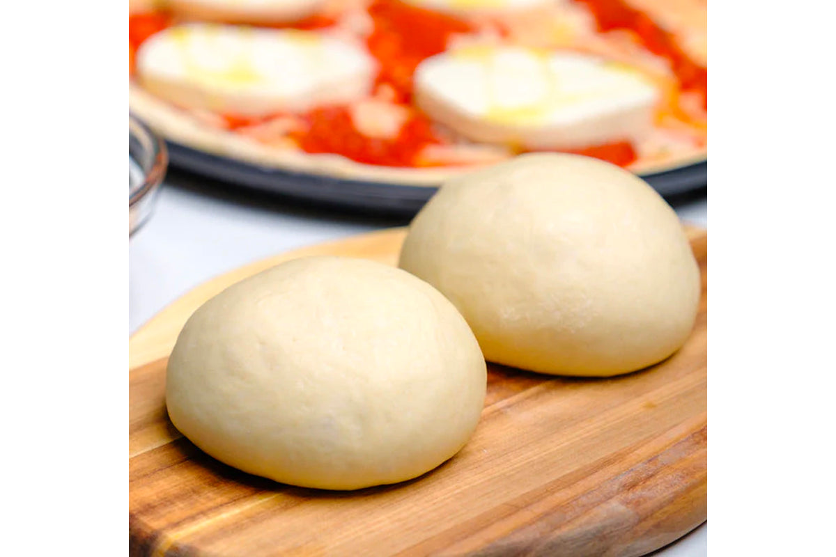 Two round pizza dough balls on a wooden board with finished pizzas in the background.