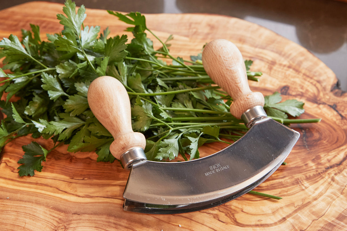 Double-bladed curved chopper with wooden handles resting on cutting board next to fresh parsley.