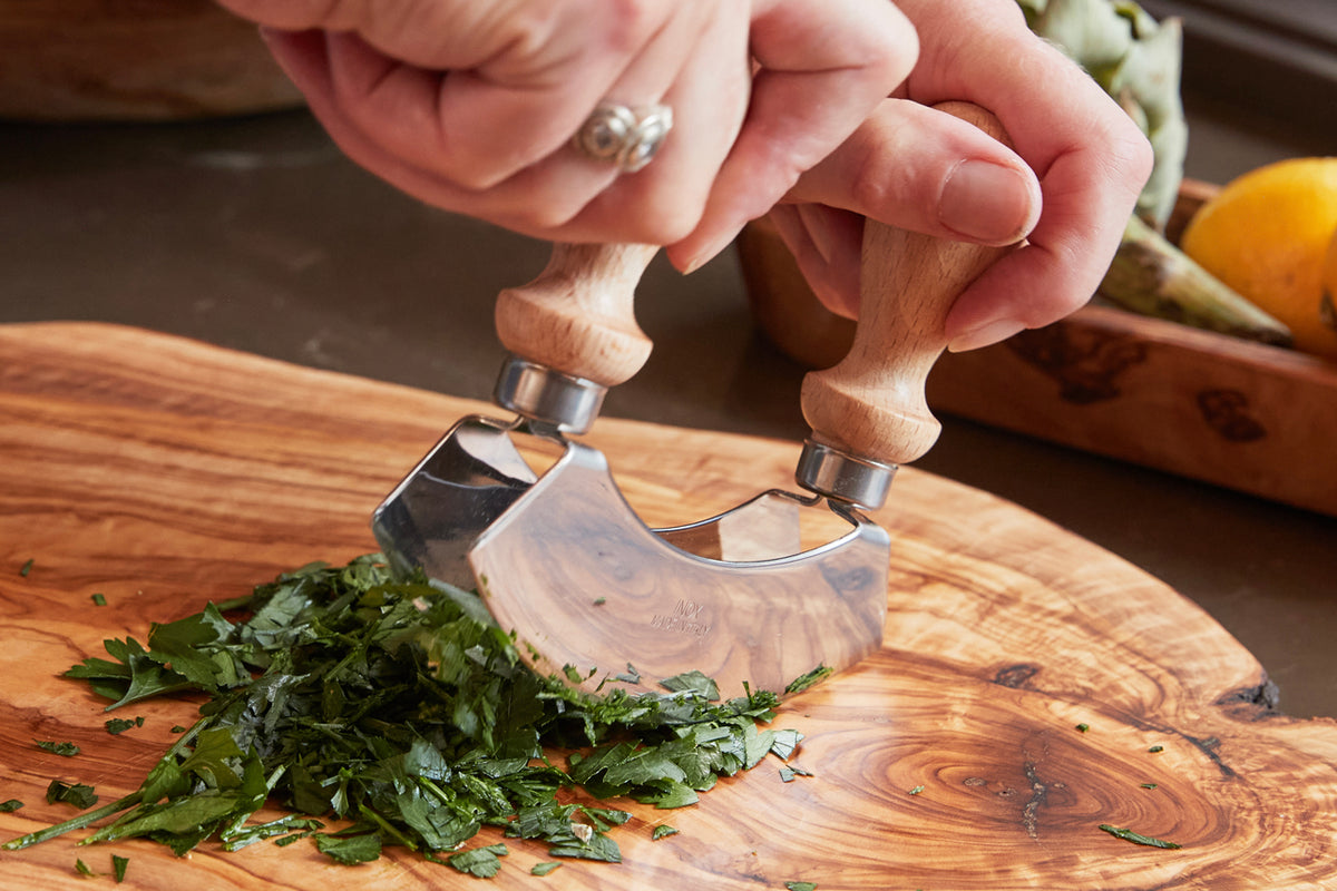 Hands holding wooden-handled double mezzaluna with curved metal blades chopping fresh herbs on wooden cutting board.