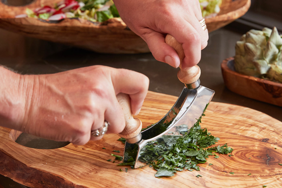 Two hands using a double-bladed curved chopper with wooden handles to mince fresh herbs on a wooden cutting board.