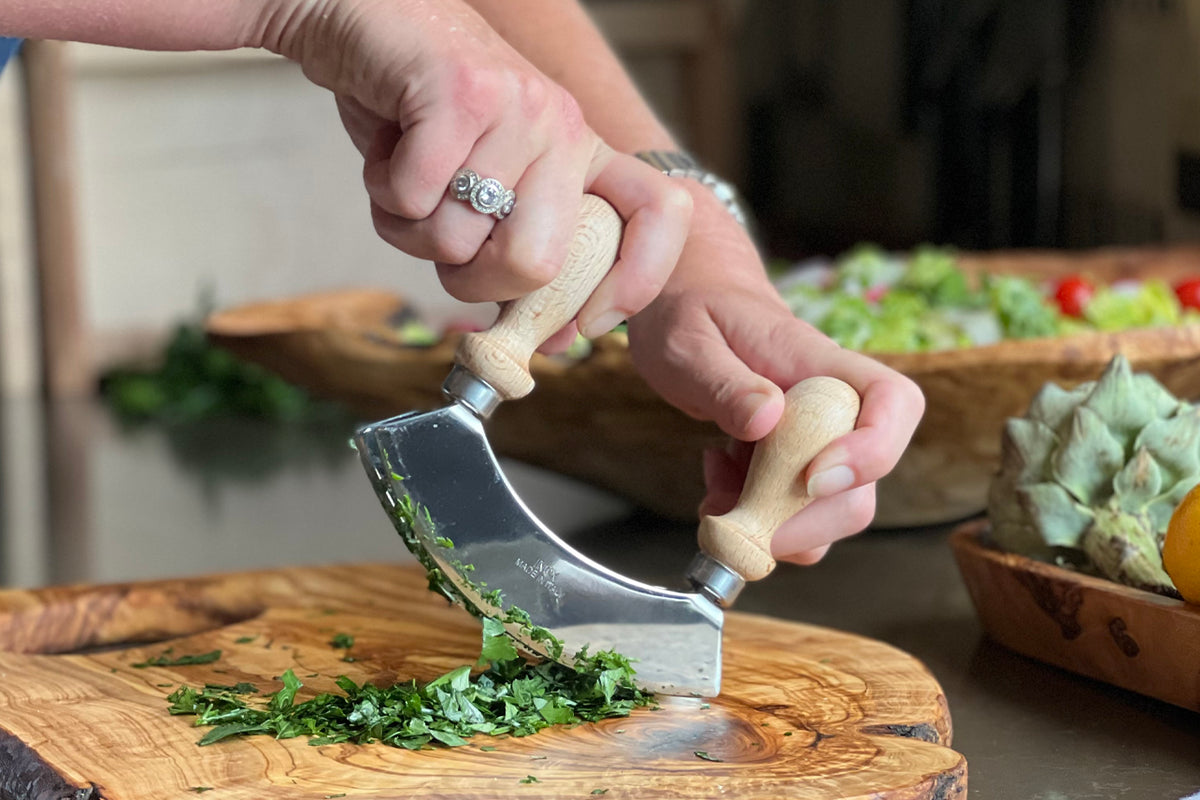 Hand holding double-bladed mezzaluna with wooden handles chopping herbs on wooden cutting board.