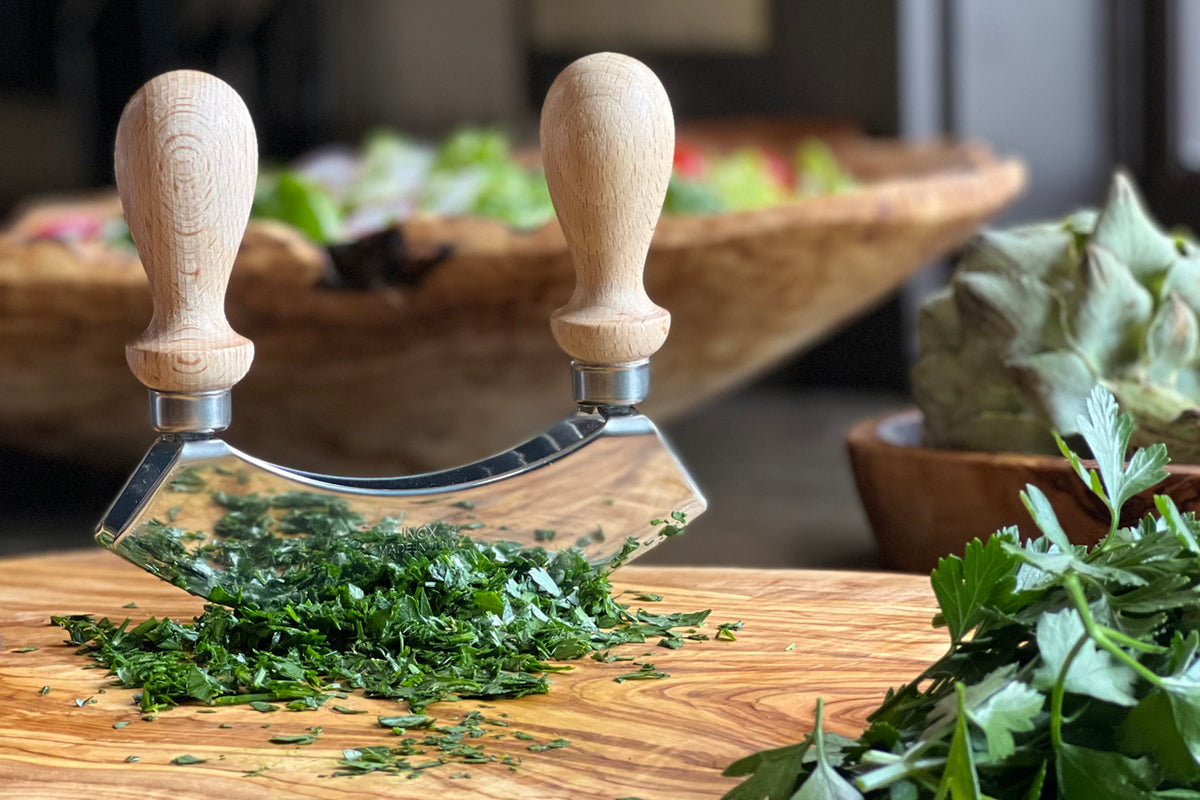 Double-bladed mezzaluna chopper with wooden handles and metal blades on cutting board with chopped herbs.