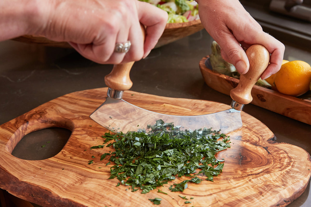 Two hands grip wooden handles of a curved metal blade chopping herbs on a wooden board with handles.