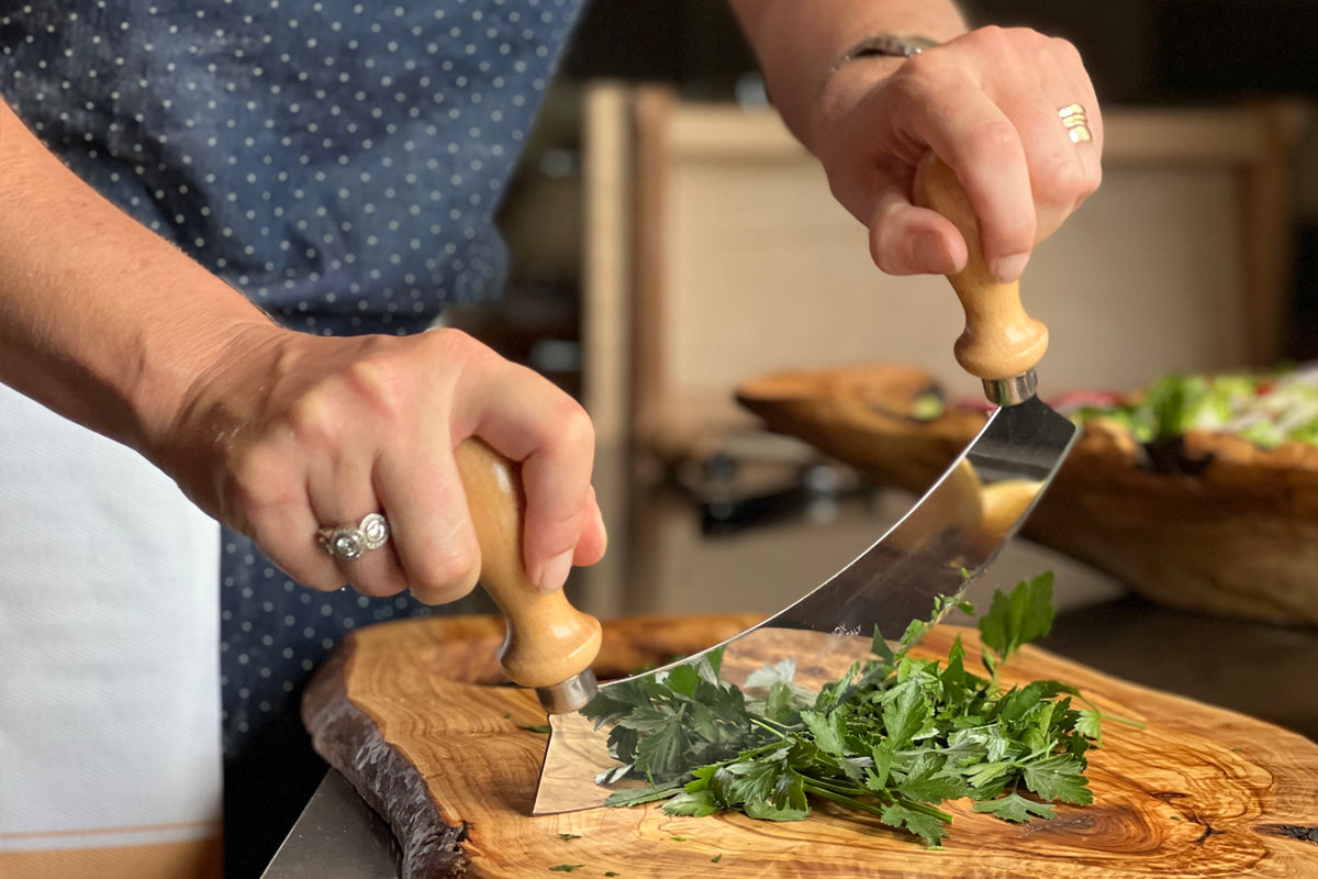 Person using mezzaluna with wooden handles to chop parsley on a wooden cutting board.