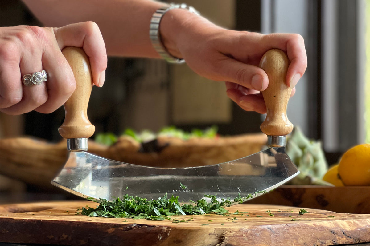 Large mezzaluna with wooden handles and curved metal blade chopping fresh herbs on cutting board.