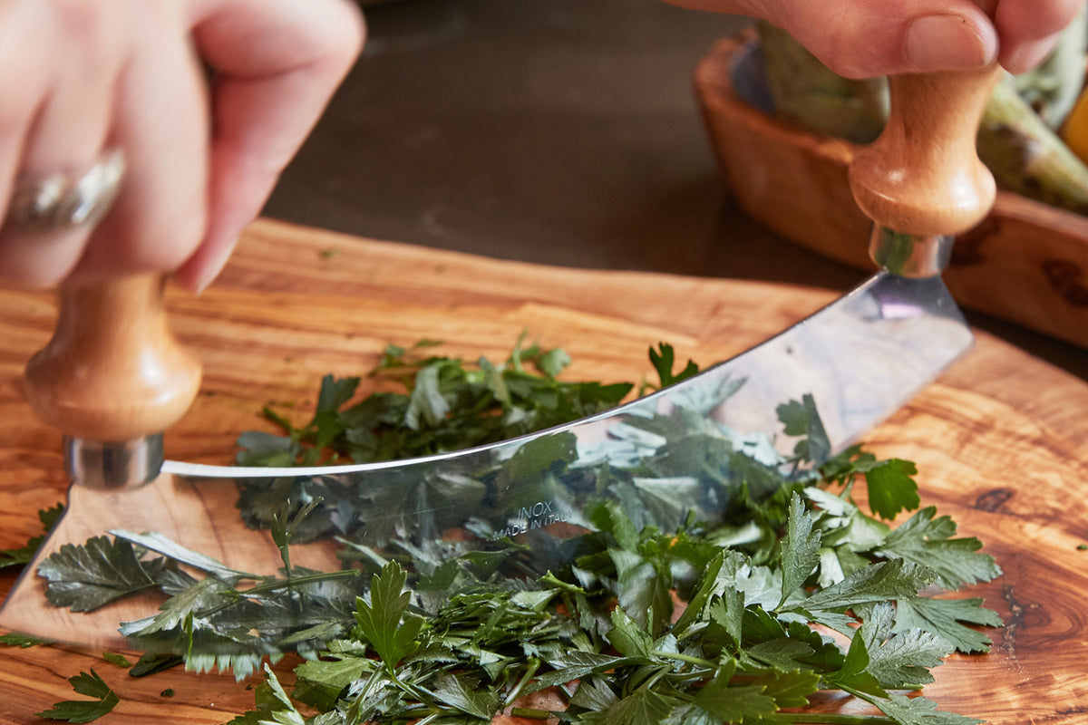Large mezzaluna with wooden handles chopping fresh parsley on a wooden cutting board.