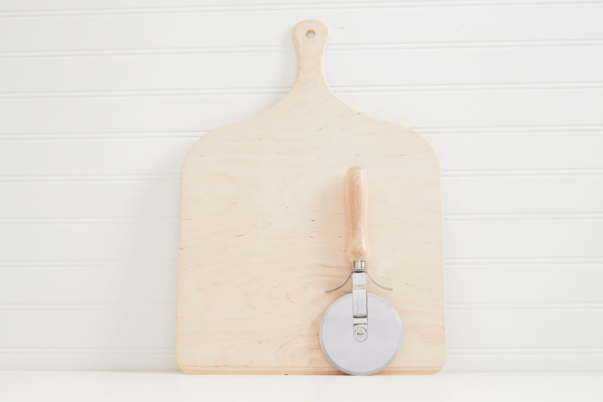 Pale wooden pizza peel with handle and metal pizza cutter wheel attached to the side.