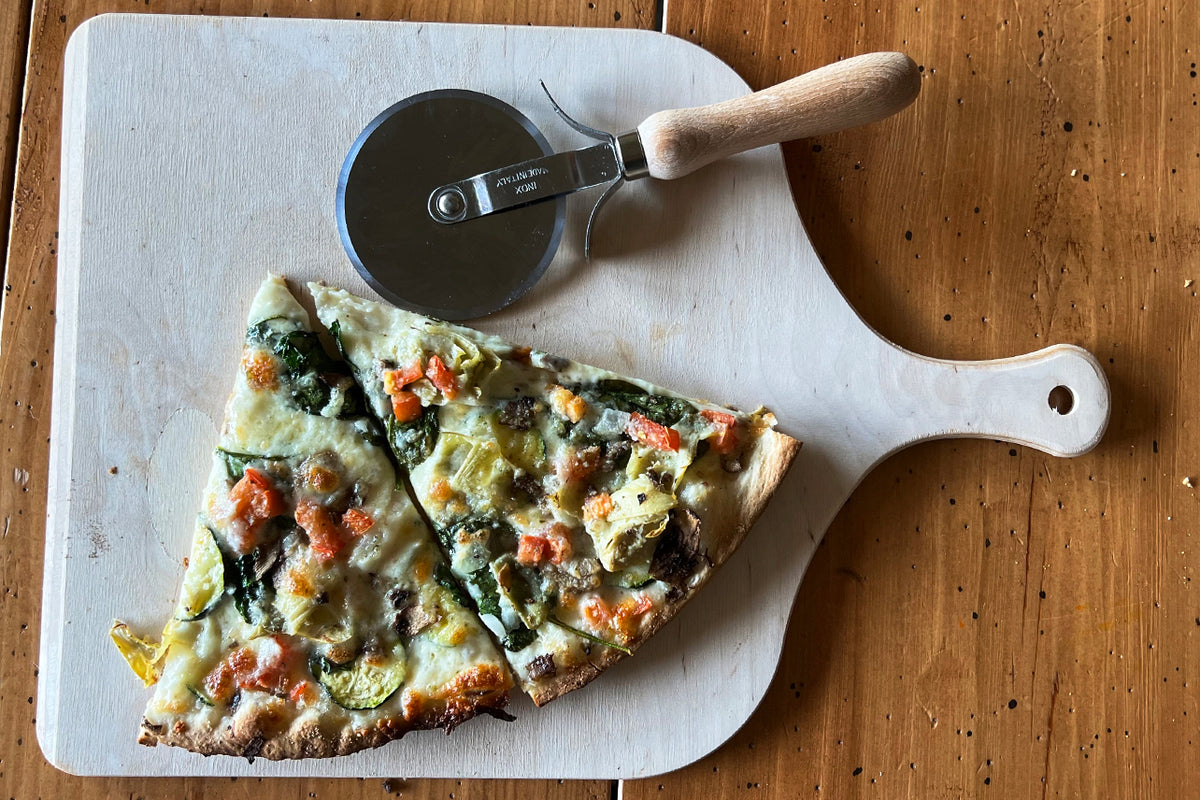 Light wood pizza peel with handle holding a slice of vegetable pizza next to a pizza cutter.