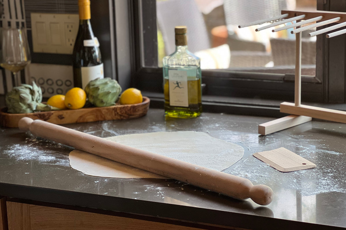 Wooden pasta rolling pin with dough scraper on floured counter surface.