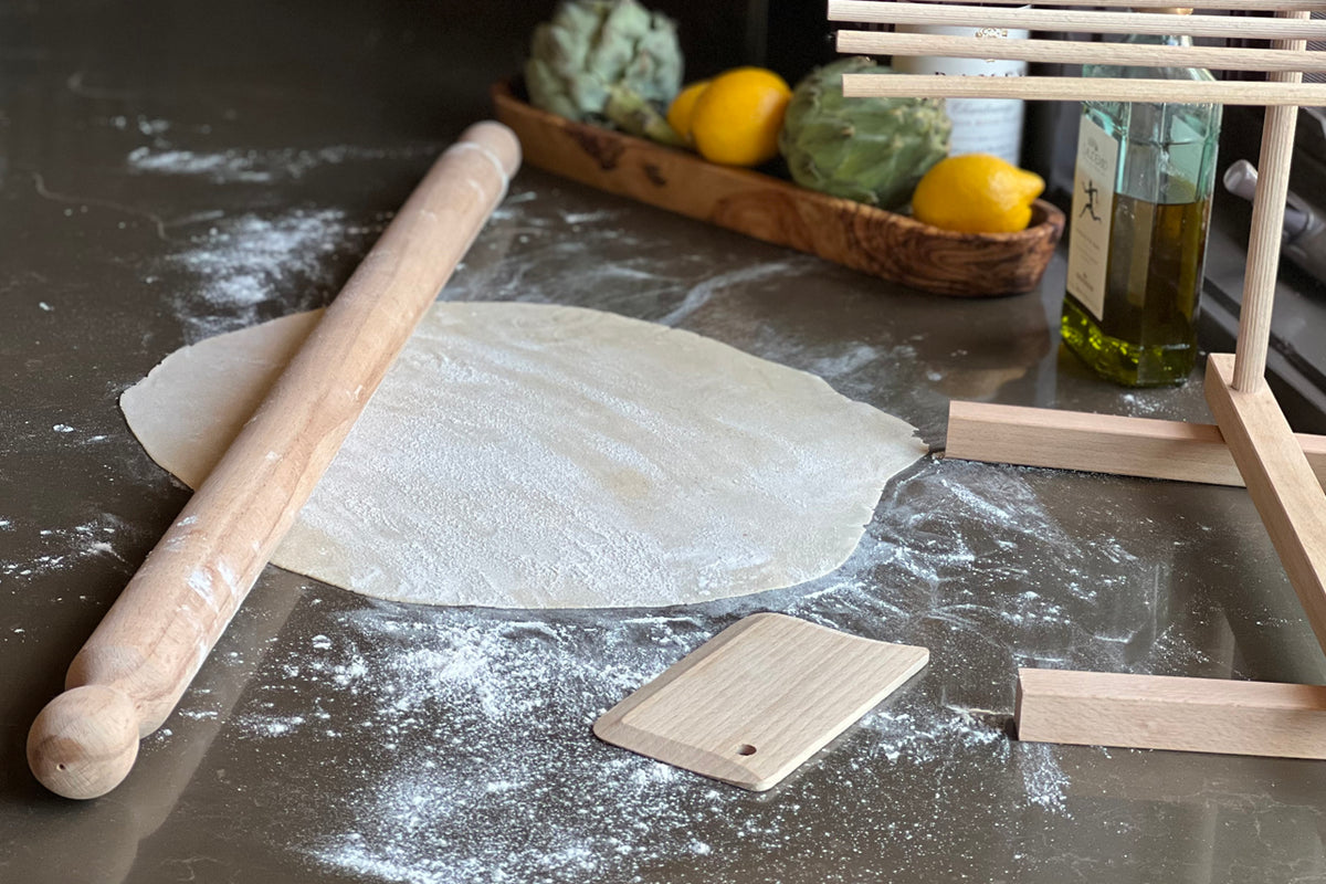 Wooden pasta roller and rectangular dough scraper on floured surface with pasta dough.