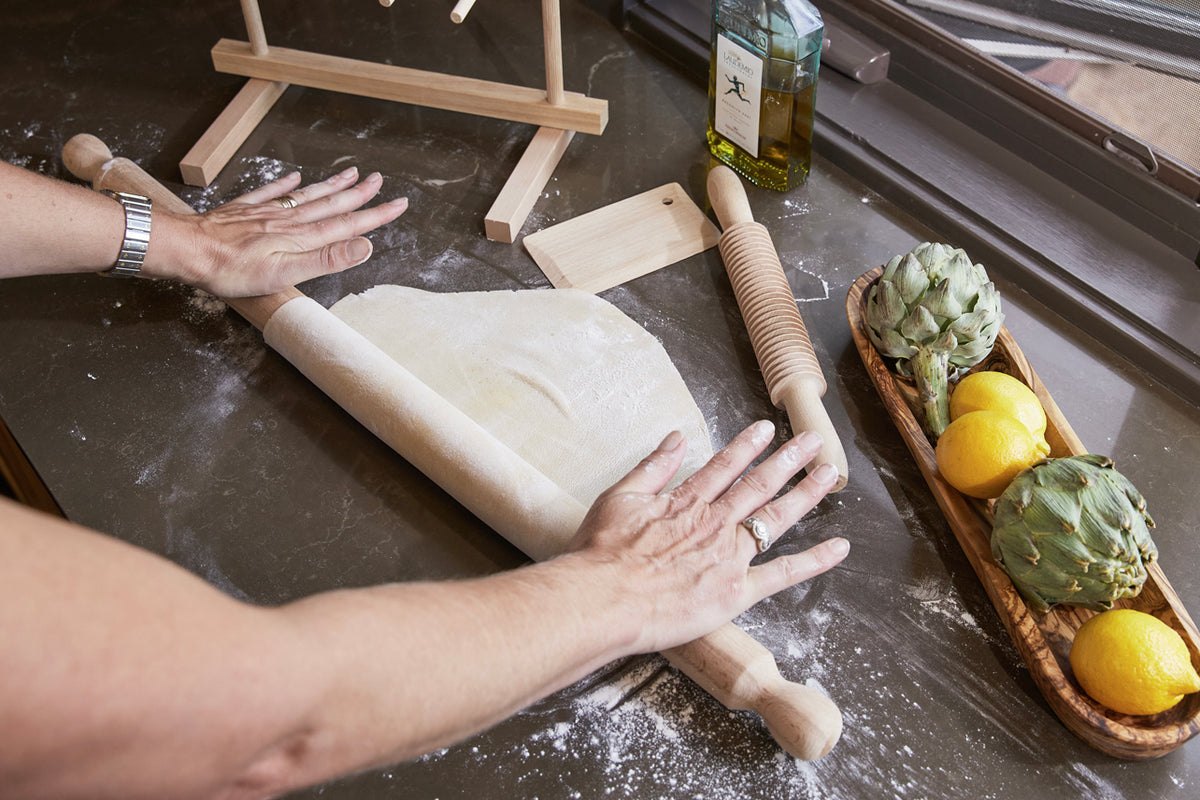 Wooden mattarello rolling pin with textured surface and dough scraper being used to flatten pasta dough on dark work surface.