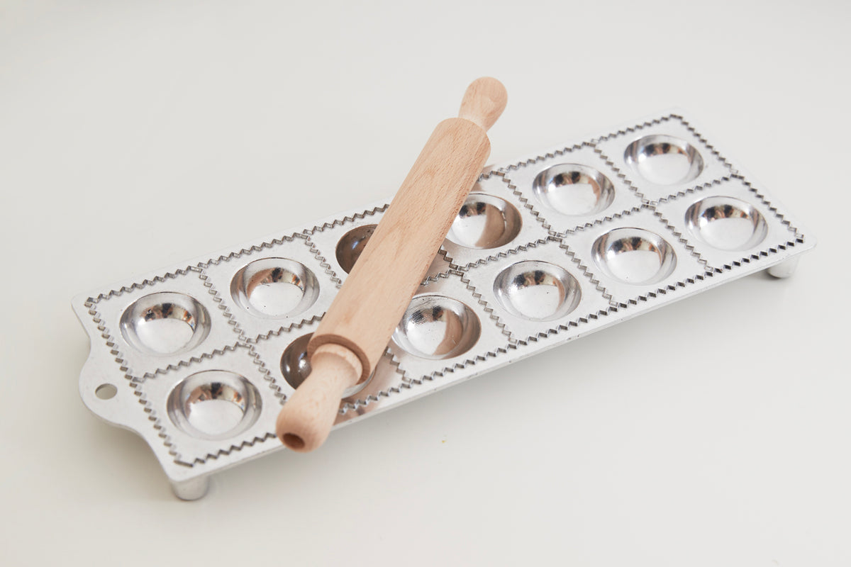 White metal ravioli mold with 12 hemispherical cavities and wooden roller with handle positioned on top.