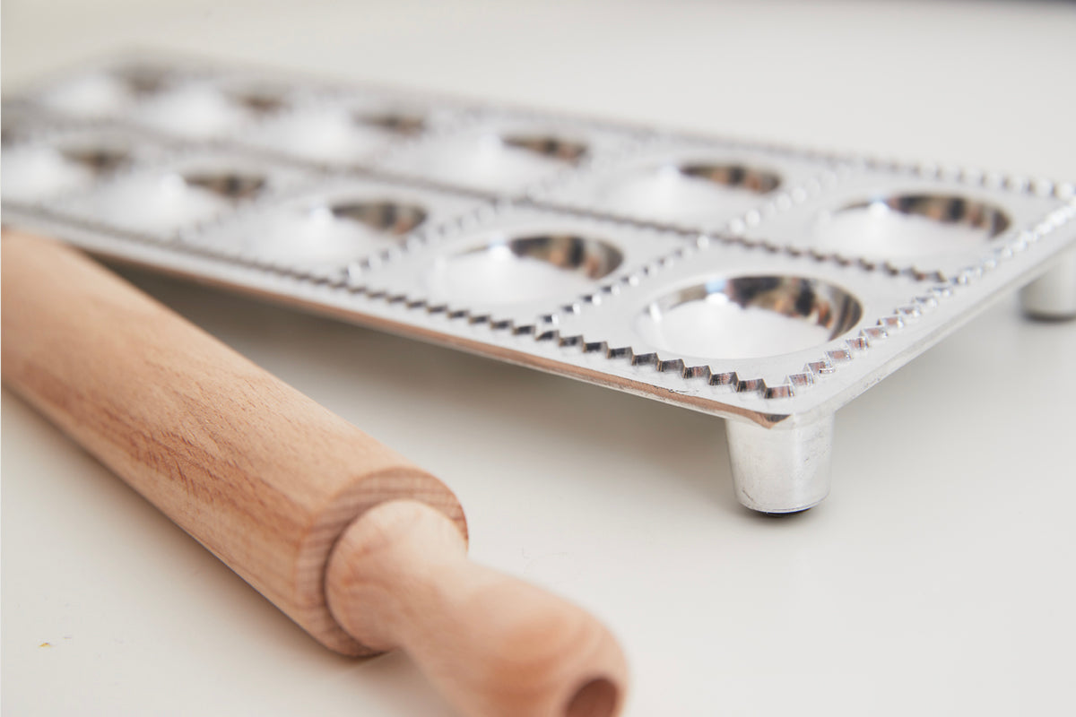 Wooden rolling pin next to white metal ravioli mold with 12 semicircular cavities and crimped edges.