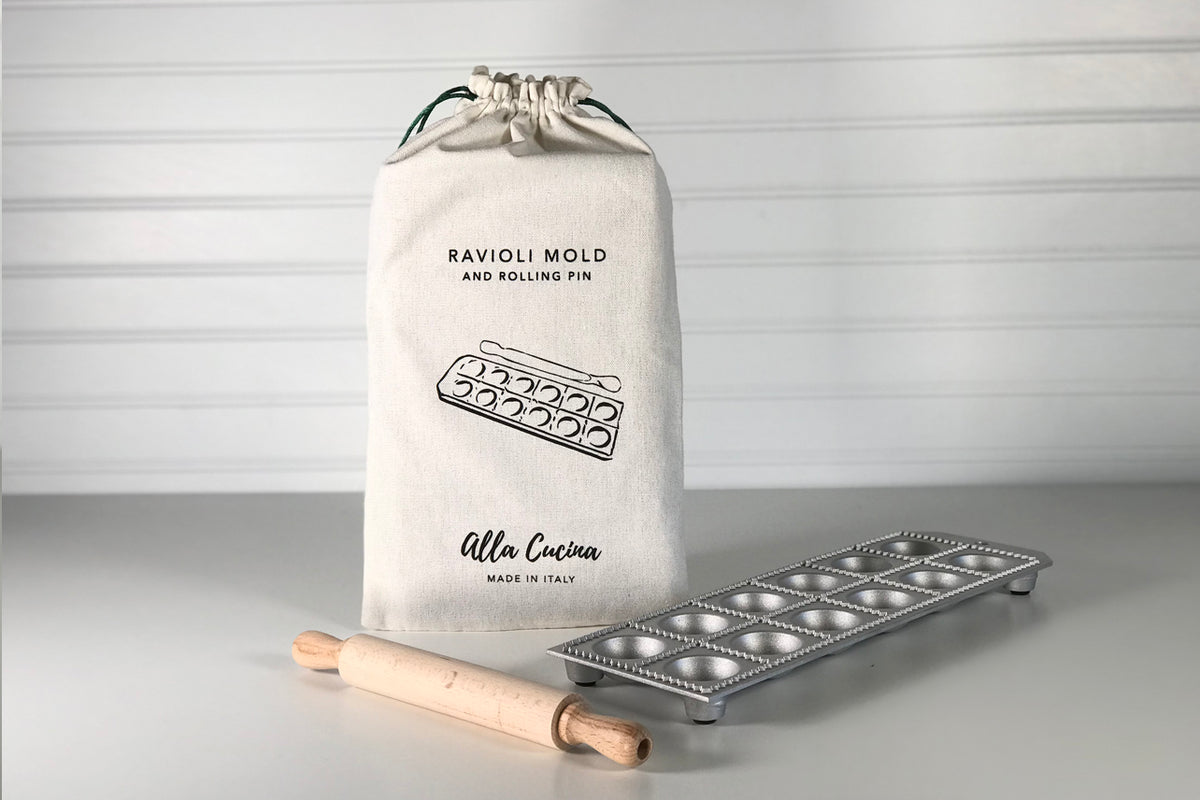 Cream canvas drawstring bag with ravioli mold and rolling pin, metal mold with wooden handles beside bag.